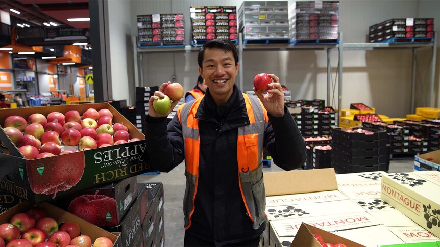 Thanh Truong, aka the Fruit Nerd, in a warehouse packed with boxes of fruit, holding a variety of apples.