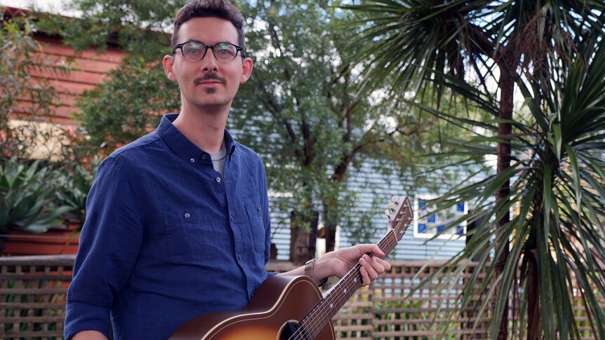 A man holding a guitar outside standing in front of some trees.