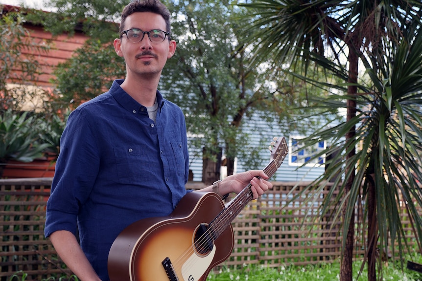 A man holding a guitar outside standing in front of some trees.