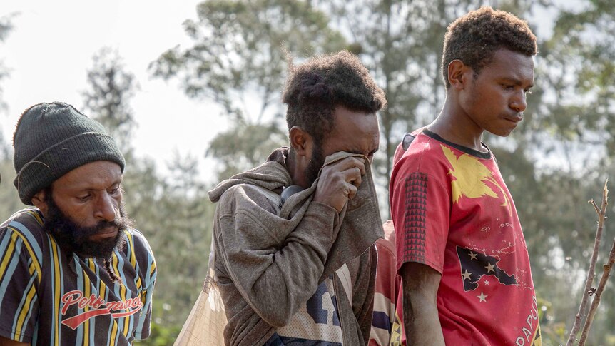 A man wipes tears away with his jacket, flanked by two other men looking downcast.