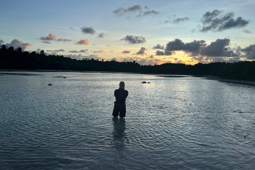 A woman, sihouetted by the setting sun, stands knee-deep in water.
