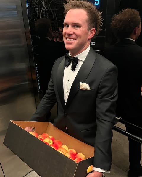 A man wearing a tuxedo holds a box of apples, smiling.