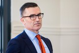 A man in a suit with an orange tie and glasses looking towards the camera with a serious expression.