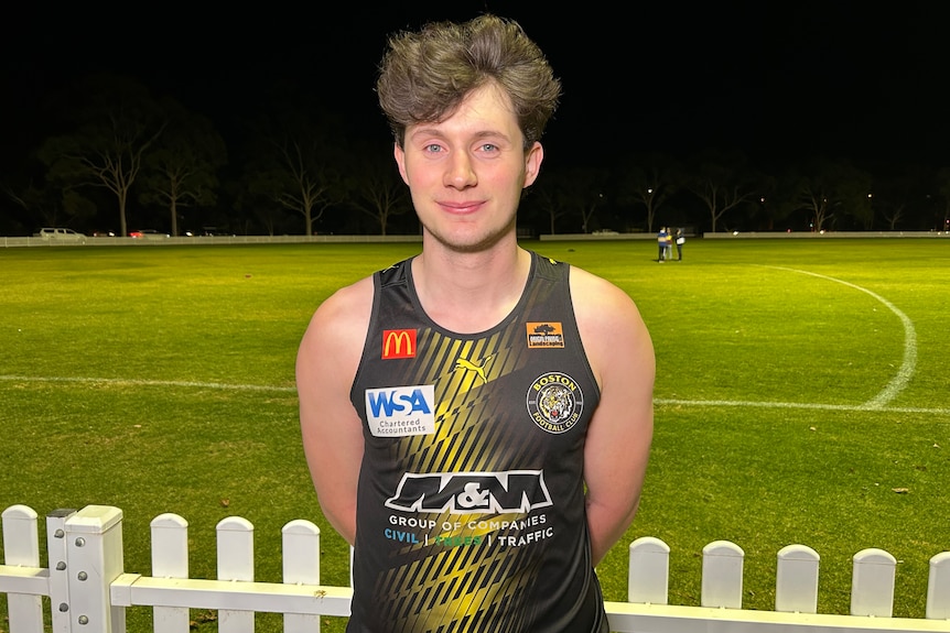 A man in a black and yellow footy jersey next to an oval.