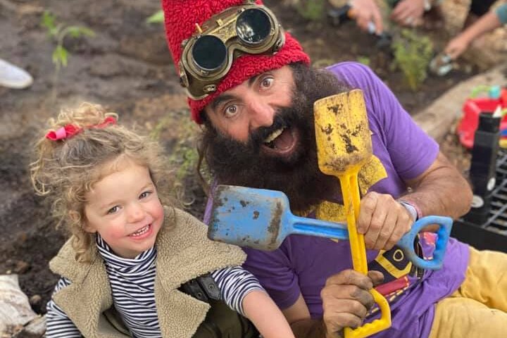 A young girl sits in the dirt next to a man with a huge beard in fancy dress holding two coloured mini shovels. 
