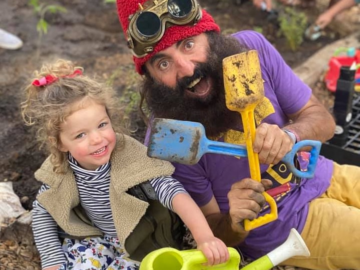 A young girl sits in the dirt next to a man with a huge beard in fancy dress holding two coloured mini shovels. 