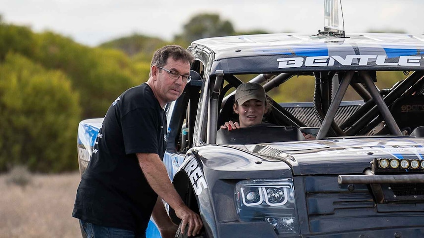 A man leans against the outside of a racing buggy, next to a young woman sitting in the driver's seat.