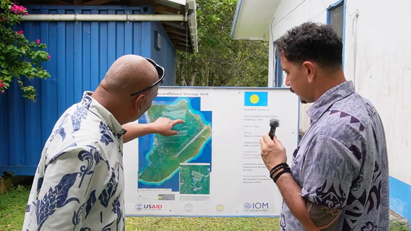 Two men stand in front of a map of Angaur.