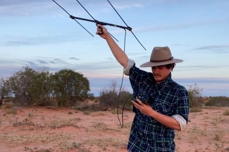 Man wearing shirt and Akubra hat stands in the desert holding a piece of equipment in the air.