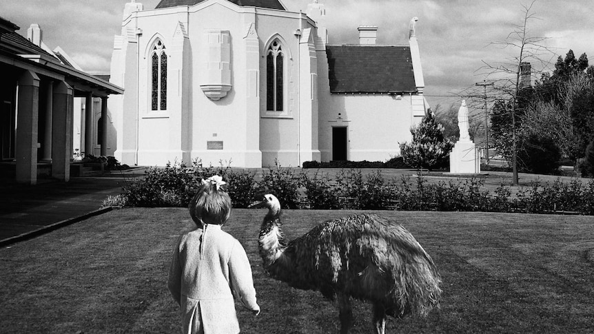 A young child and an emu stand next to each other on the lawn in front of a church which has eagles in place of crosses