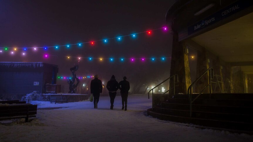 A trio of snowgoers stroll under fairy lights through the Mt Buller village at night.