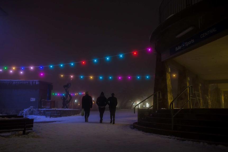A trio of snowgoers stroll under fairy lights through the Mt Buller village at night.