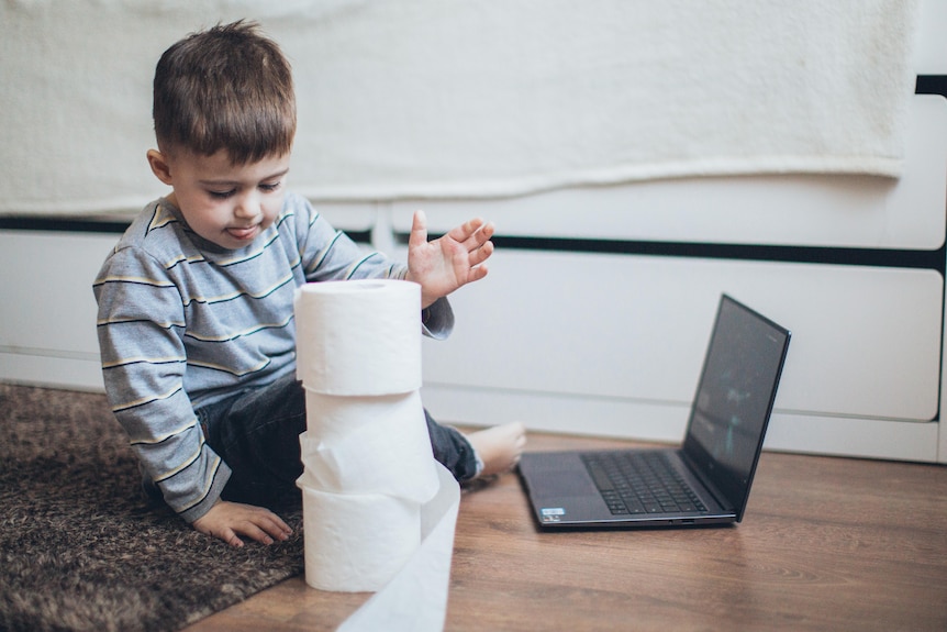 A toddler pokes his tongue out and prepares to hit a stack of rolls of toilet paper, sitting on the ground with a laptop.