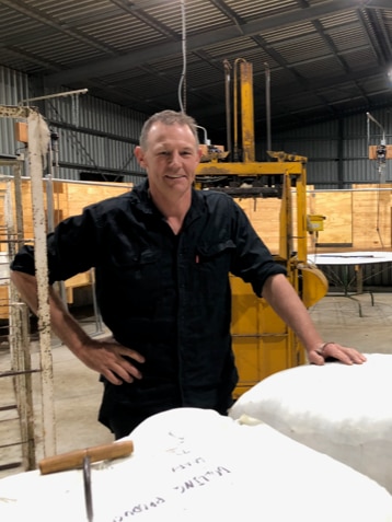 A man standing in a woolshed behind wool bales.