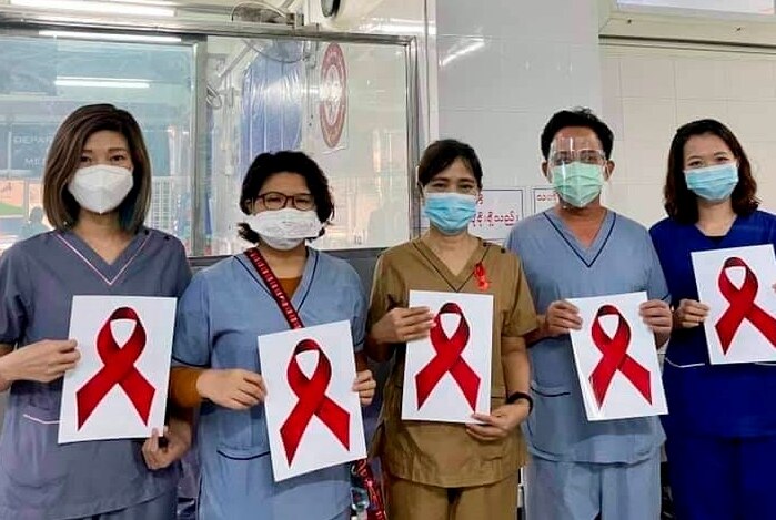 Hospital workers in scrubs and face masks hold up sign with red ribbon.