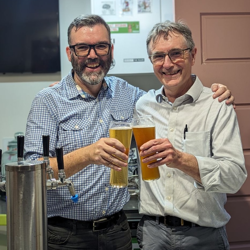 two men, one with spectacles and beard and other with grey hair, each holding a beer together