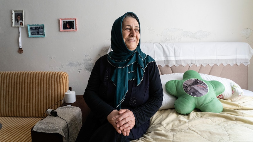 An older woman in a deep turquoise hijab sits on a bed with her hands folded on her lap, smiling