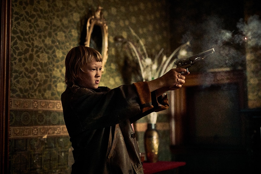 A boy with blonde cropped hair in oversized jacket stands in dimly lit 1870s interior holding smoking pistol with two hands.