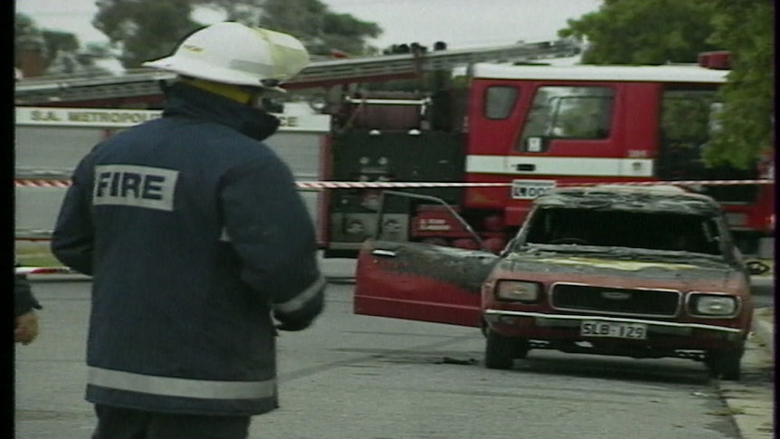 A firefighter has his back to the camera, a burnt out car can be seen in the background.