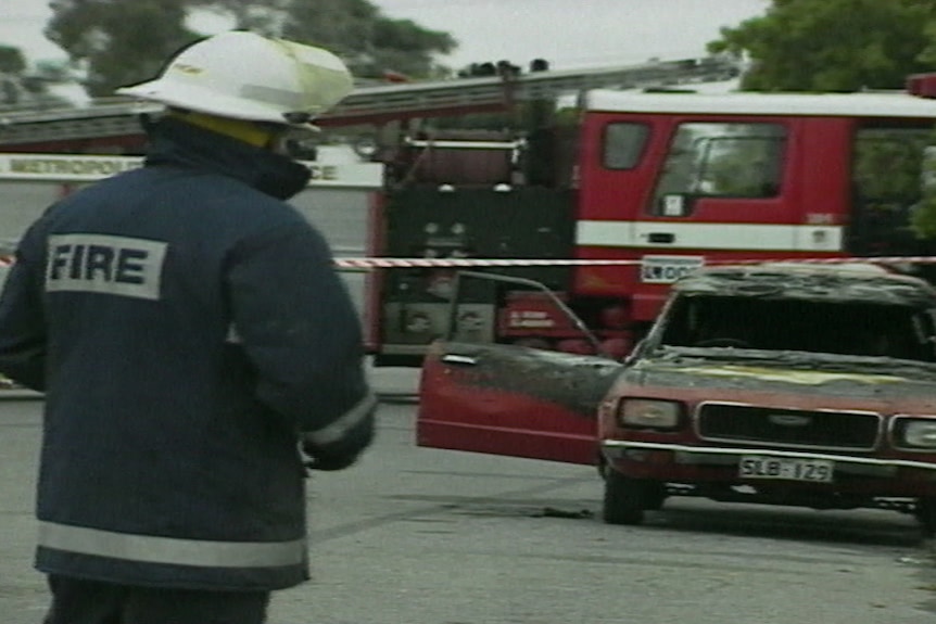 A firefighter has his back to the camera, a burnt out car can be seen in the background.