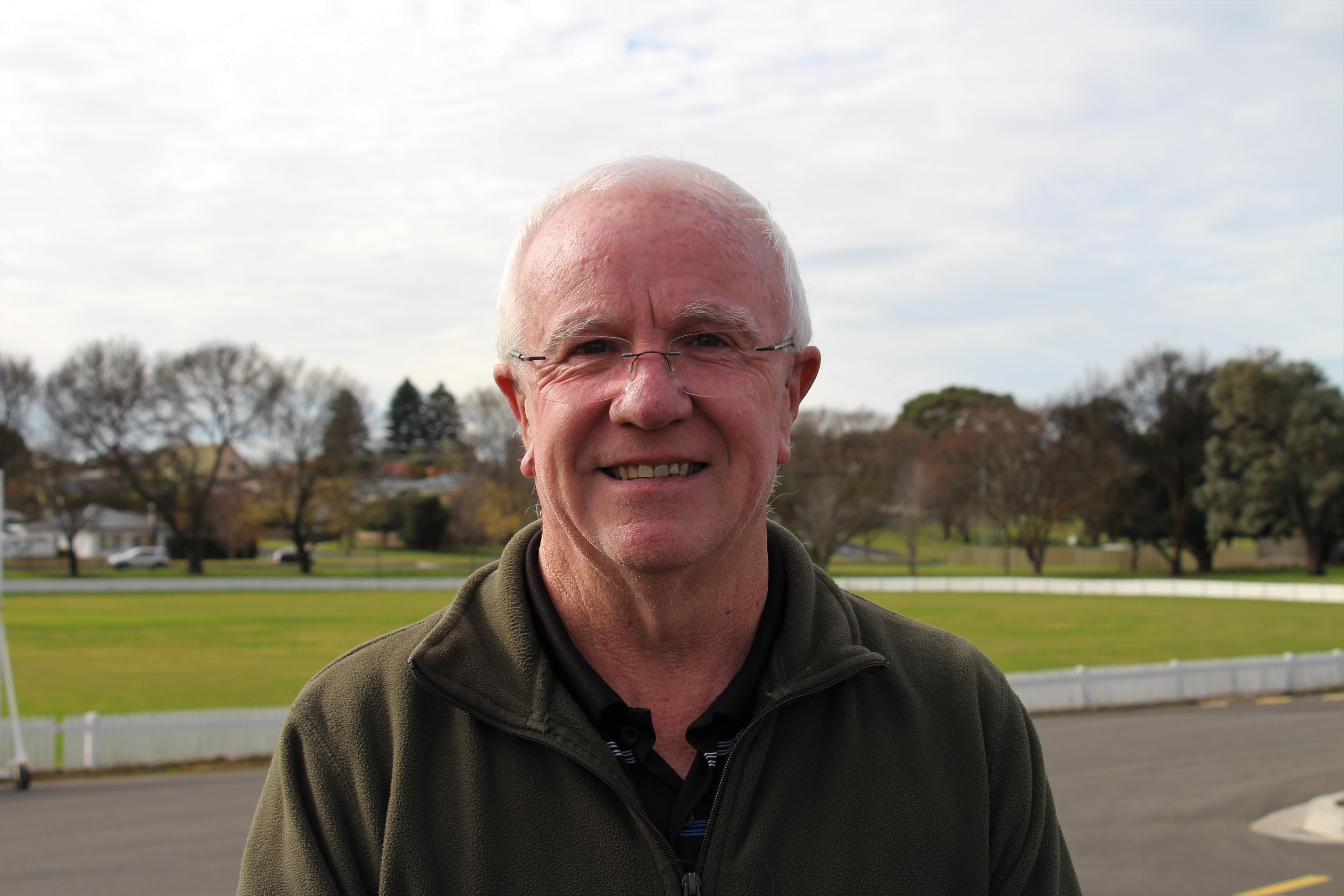 A white-haired man with glasses smiles in front of a green oval.