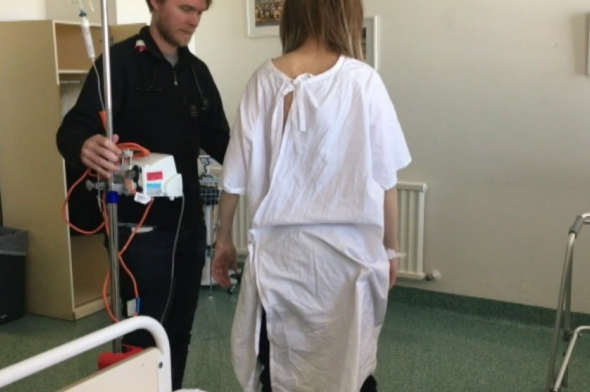 A woman in a hospital gown leans on a nurse for support in a hospital room