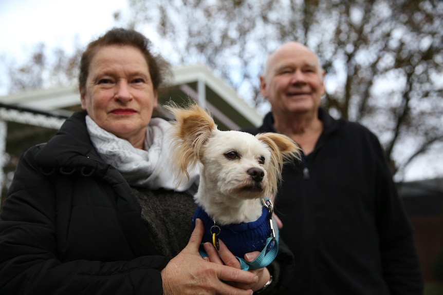 A couple hold their small white dog on a grey day. 