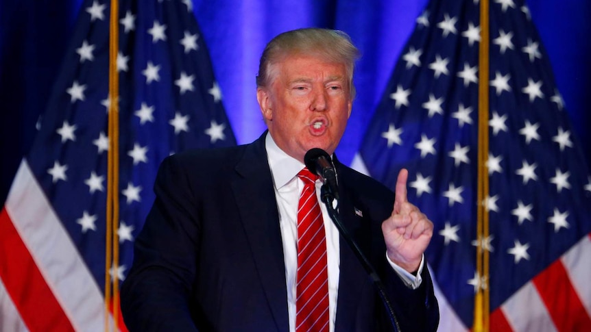 Donald Trump speaks into a microphone, pointing a finger in the air, with two American flags behind him.