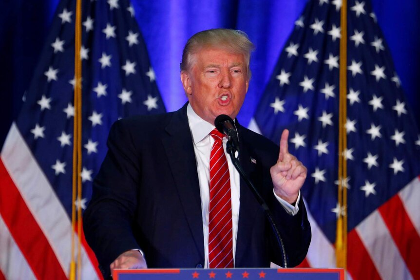 Donald Trump speaks into a microphone, pointing a finger in the air, with two American flags behind him.