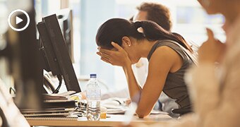 A woman sits in an office in front of a computer screen with her head down and hands covering face.