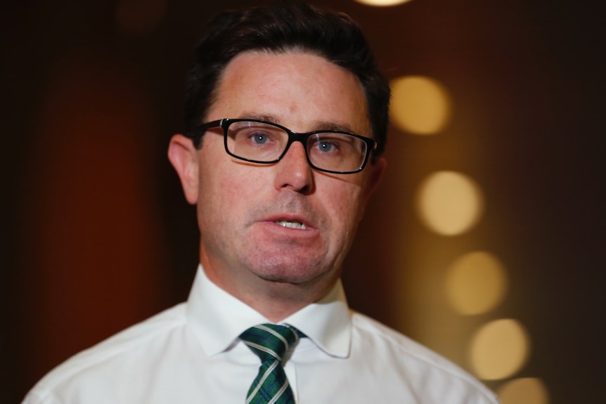 A man with brown hair and glasses wearing a white shirt and dark striped tie