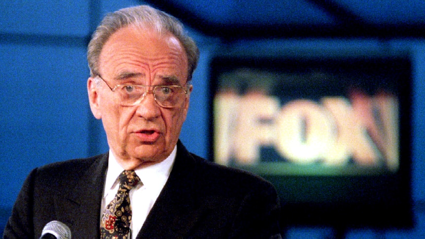 Rupert Murdoch gestures in front of a sign that reads "Fox" 