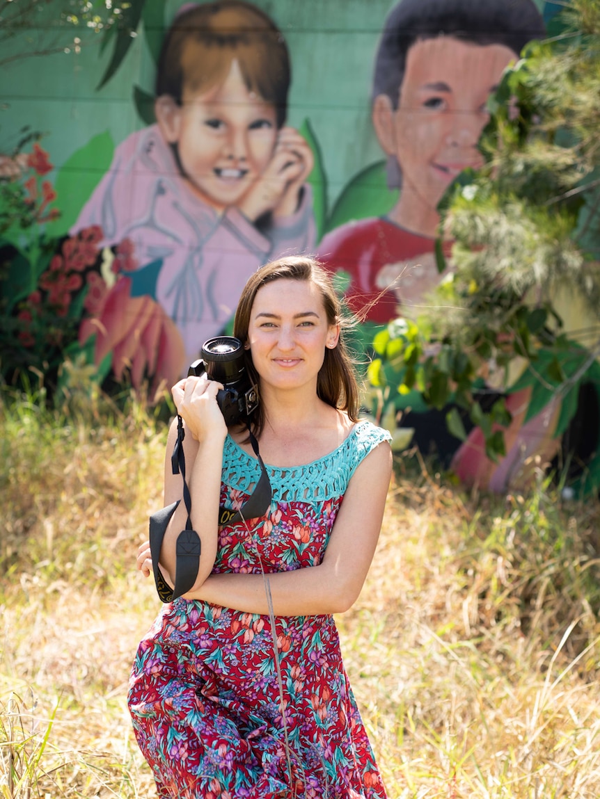 Woman stands with a camera in front of a mural of children on an highway concrete sound wall.