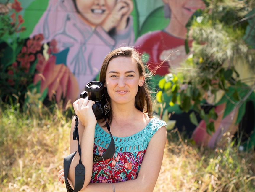 Woman stands with a camera in front of a mural of children on an highway concrete sound wall.