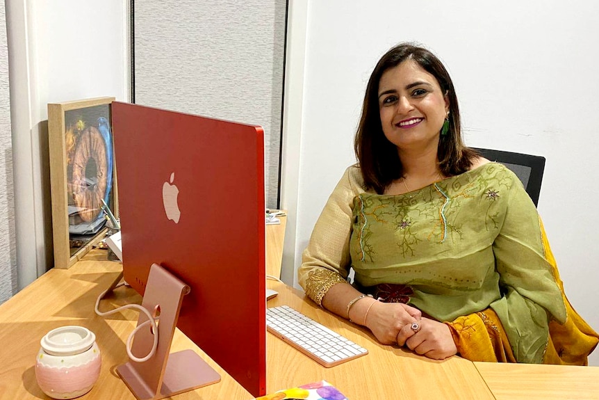 A smiling Indian woman sits at desk in front of Apple monitor, wears traditional clothes, green scarf across bust.
