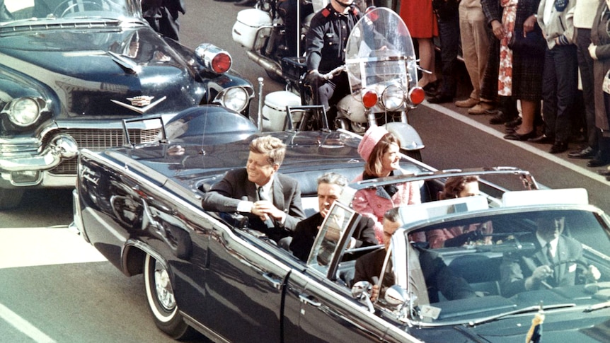 John and Jackie Kennedy ride in an open top convertible surrounded by police, onlookers stand close.