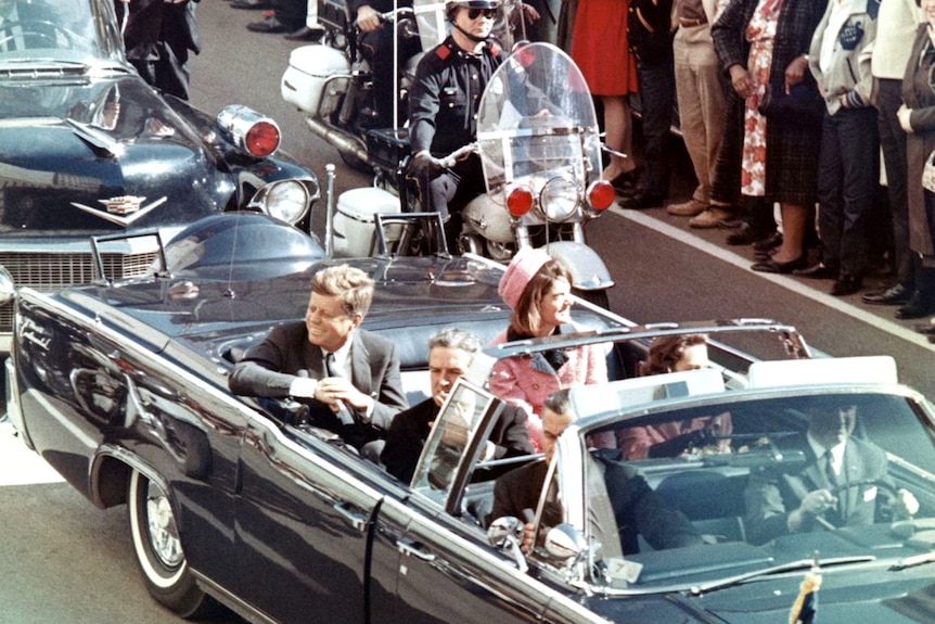 John and Jackie Kennedy ride in an open top convertible surrounded by police, onlookers stand close.