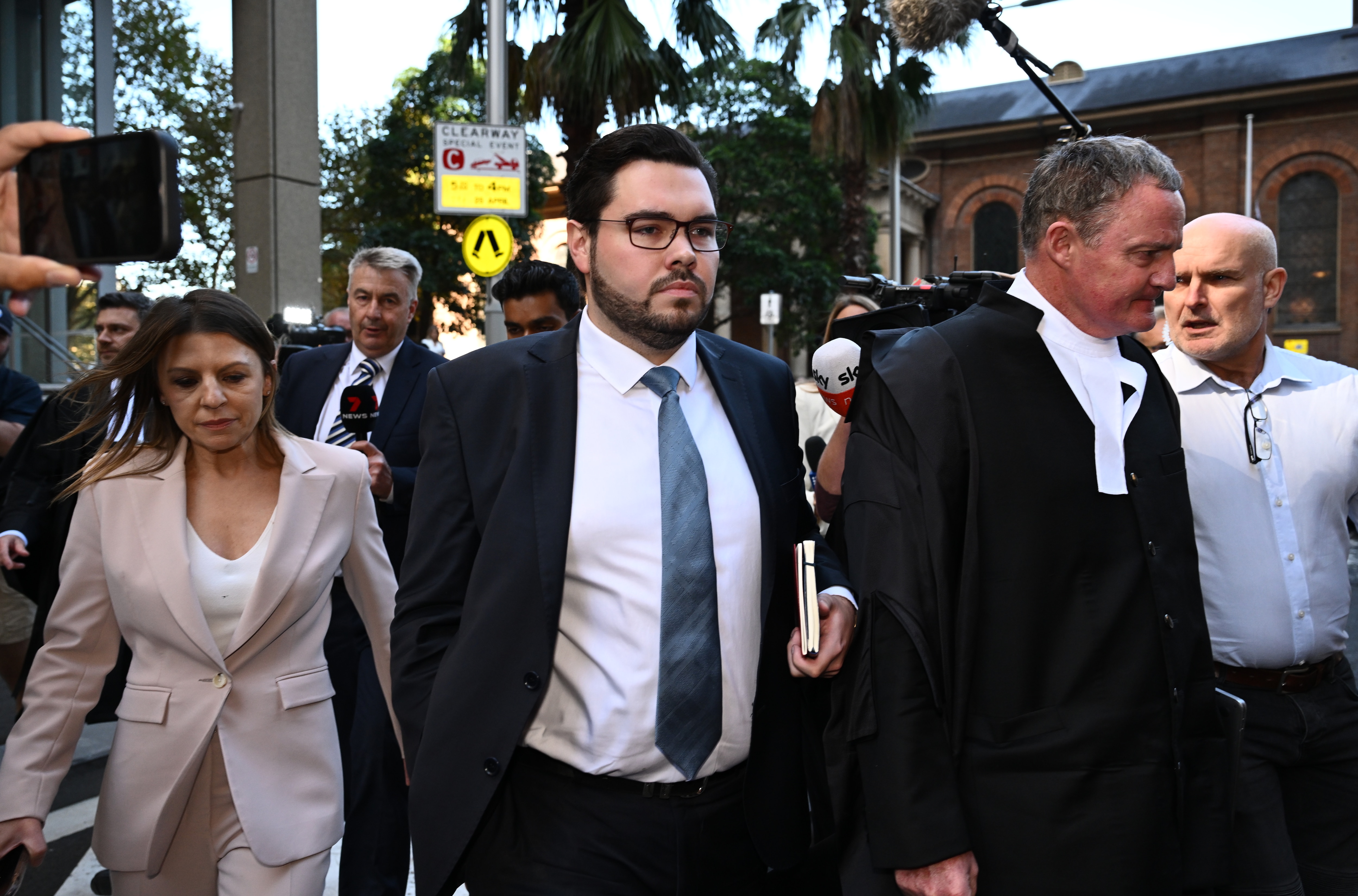 A man walks out of glass doors flanked by a lawyer and journalists.