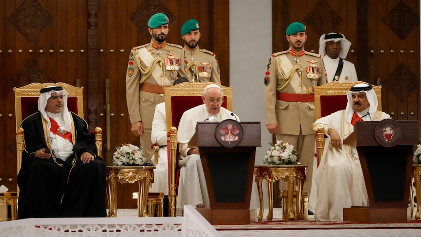 Pope Francis sits between Bahrain's king and prince in gold chairs with military officials behind. 