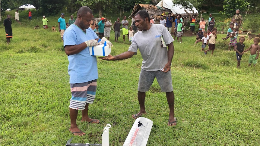 Two men hold a box in a jungle area in Vanuatu with a drone at their feet.