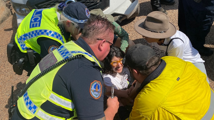 A woman sits on the ground with two police standing over her, one with handcuffs, as three other people hold her 