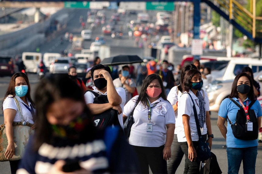 A group of people wearing face masks carry shoulder bags as they stand next to a large, busy road.