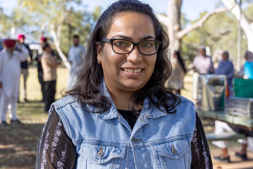 A woman in glasses looks at the camera smiling at an Indian community outdoor gathering. 