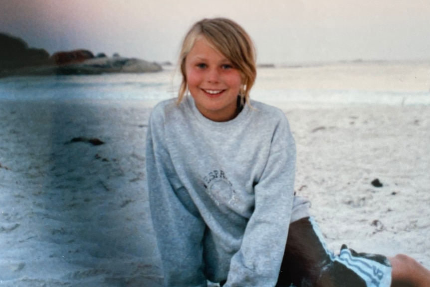 A young blonde girl sits on the sand at the beach and she wears a blue sweater