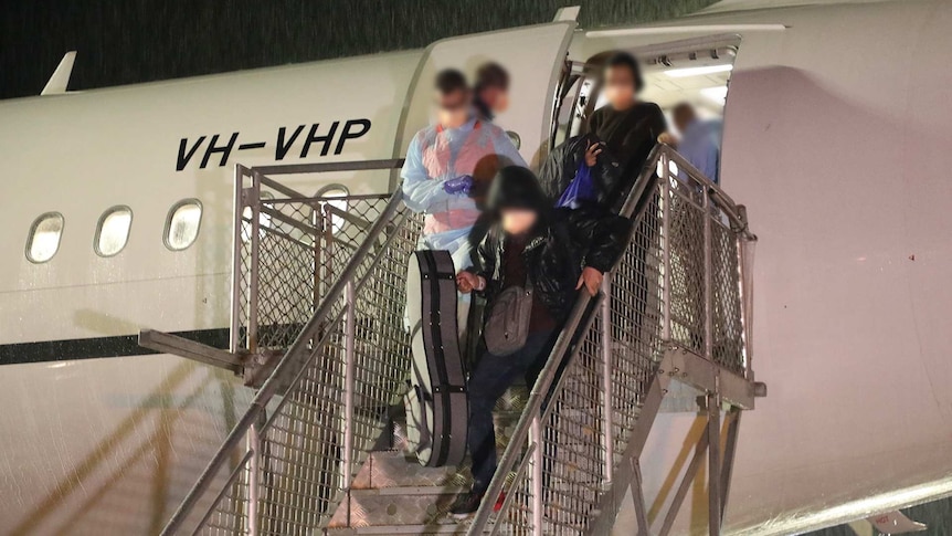 Passengers with their faces blurred disembark a plane at night.