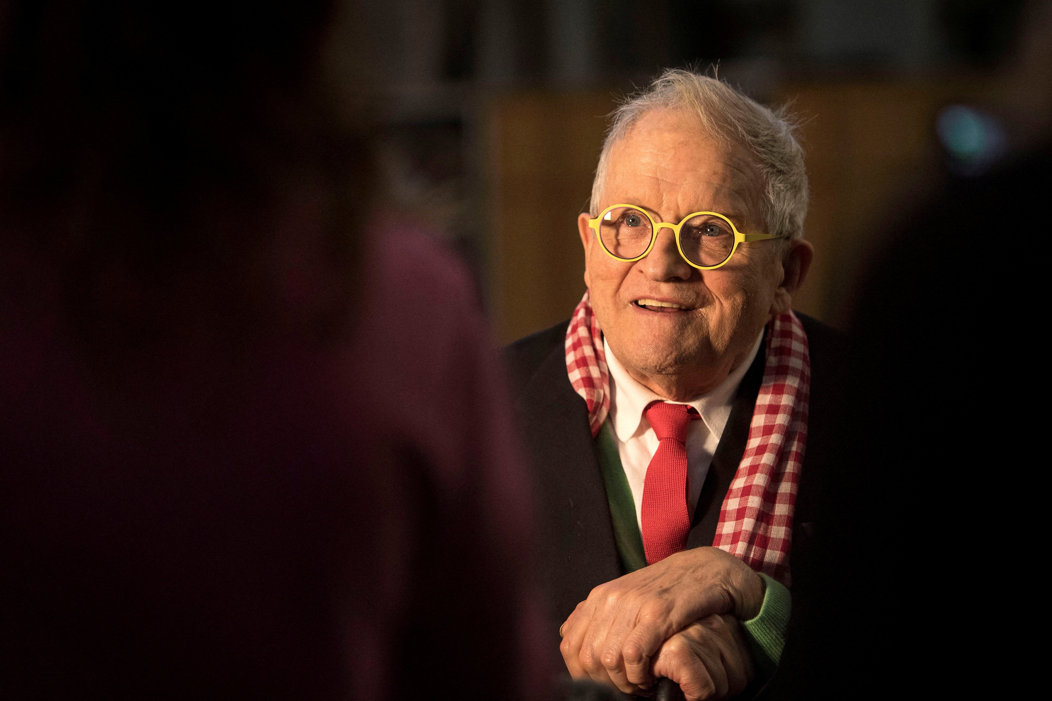 An elderly man in white glasses and red and white scarf smiles in the evening