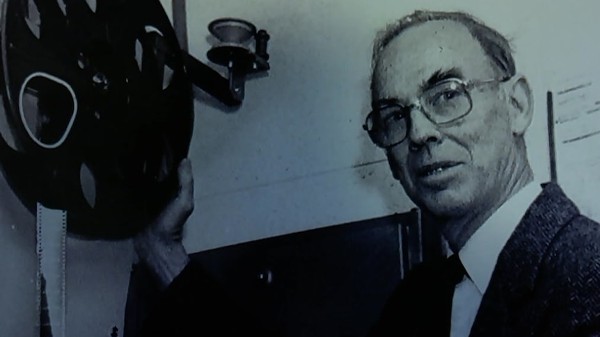 An old photo of Peter Smith wearing glasses and holding a film projector