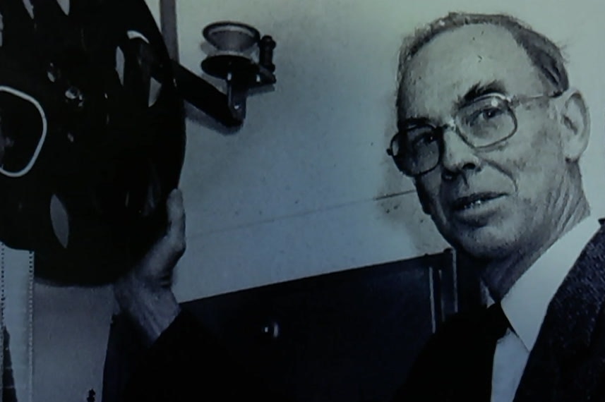 An old photo of Peter Smith wearing glasses and holding a film projector