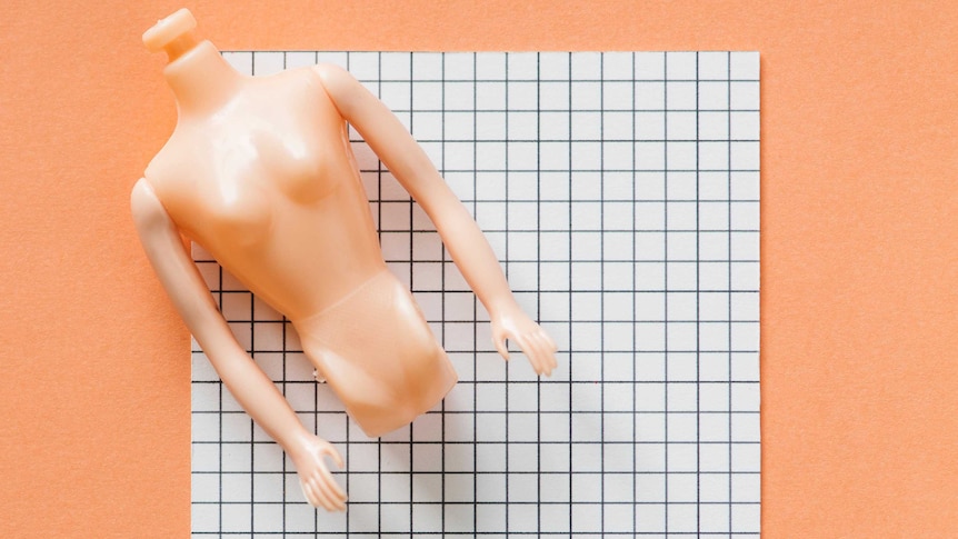 A plastic, headless doll torso sits on a piece of cardboard on a peach-coloured background.