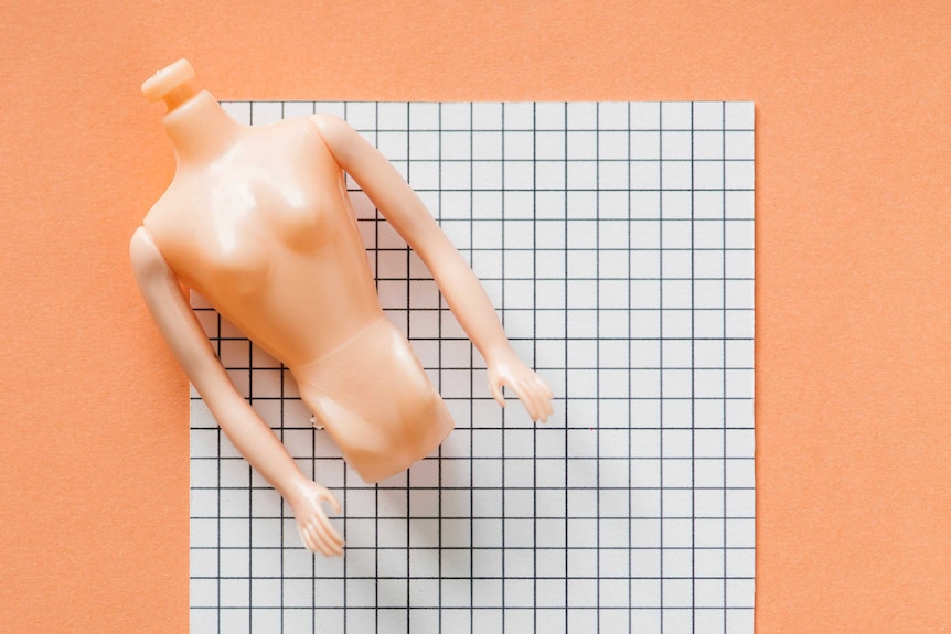 A plastic, headless doll torso sits on a piece of cardboard on a peach-coloured background.